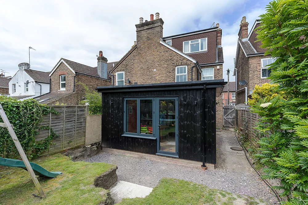 Outside rear view of kitchen extension with timber cladding and dark blue window and door frames.
