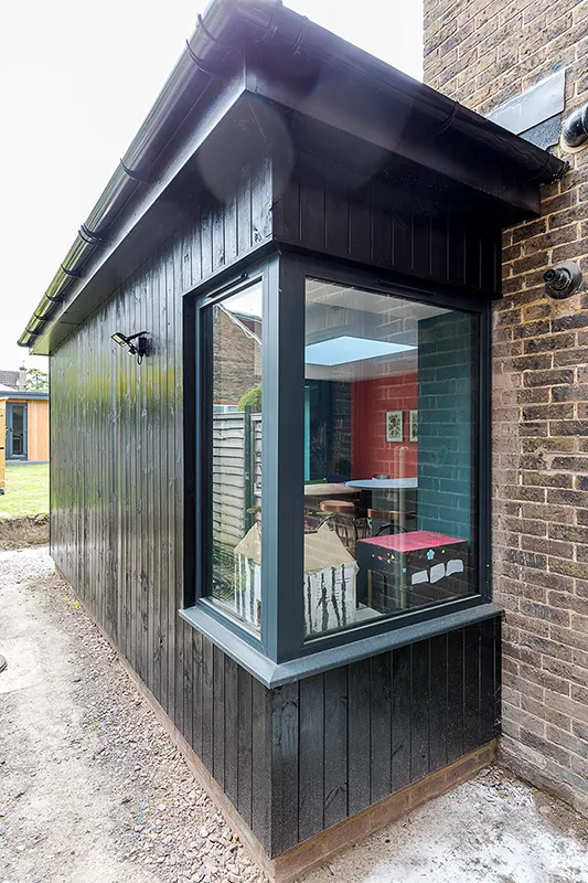 Outside rear view of kitchen extension with timber cladding and corner window with dark blue frame.